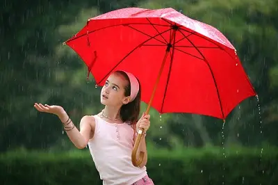 A young girl holding a red umbrella and standing in the rain, with a backdrop of green foliage.