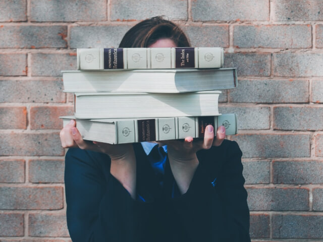 The image shows a person's hands holding a stack of books in front of their face, obscuring their identity.