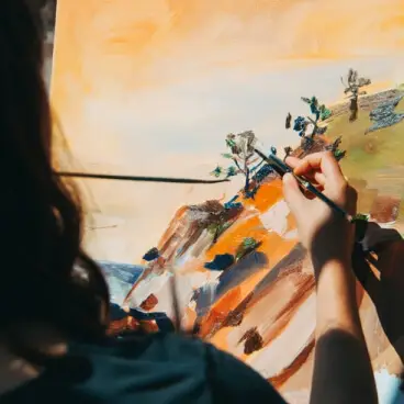 A person's hands holding a paintbrush and painting on a canvas, creating an abstract landscape